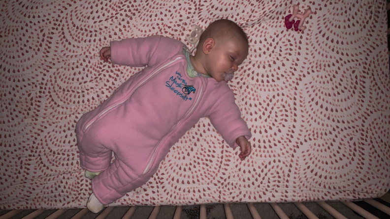 An infant lays in a crib wearing a pink sleepsuit.