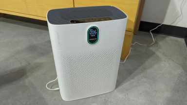 The white air purifier sits on a cement floor, its front face has perforations, welcoming smoke.