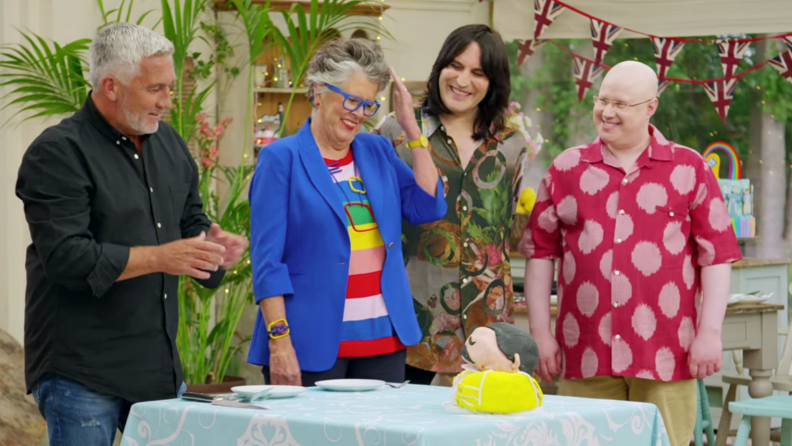 A still from The Great British Baking Show featuring Noel Fielding, Matt Lucas, Prue, and Paul Hollywood judging a cake.
