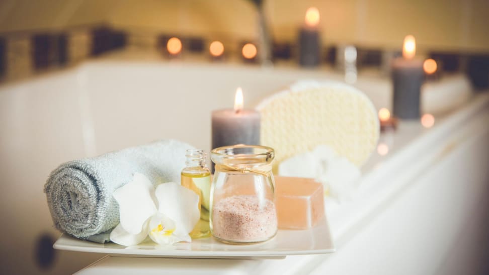 A bathtub with lit candles surrounding and a bath tray with a towel, bath salts, and a large candle