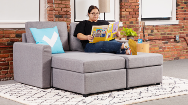 A person reading a book on a modular couch in front of brick wall indoors with their feet crossed at the ankle.