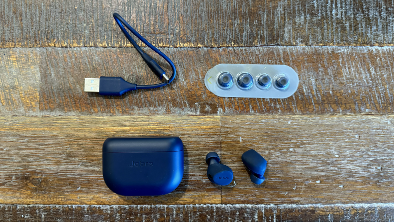 The Jabra Elite 8 Active on a wooden table next to their charging case, extra ear tips, and USB-C charging cable.
