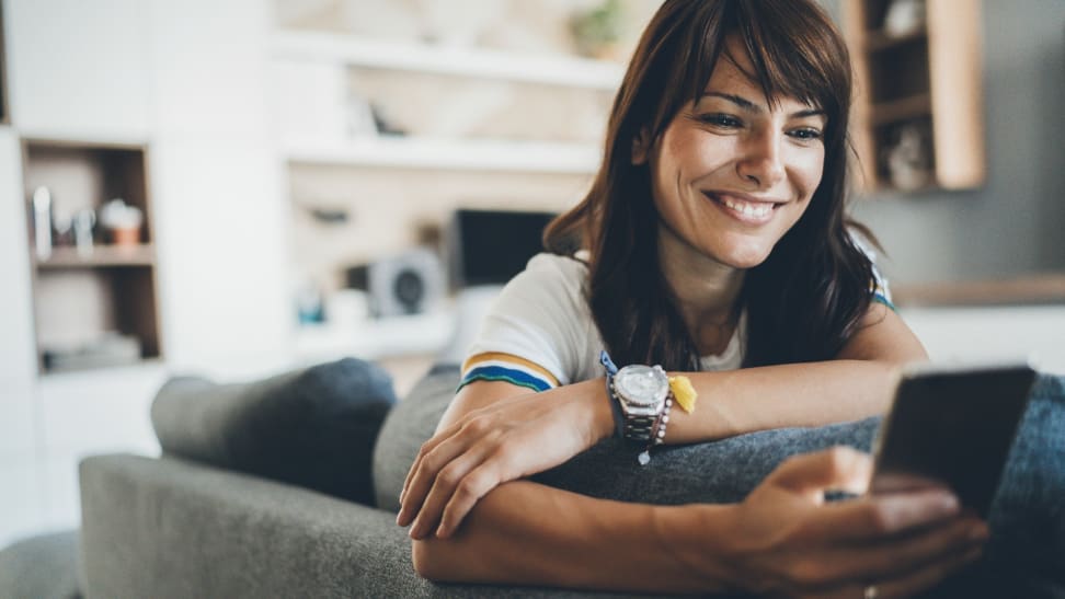 Person smiling and looking at smart phone while leaning across the back of a couch