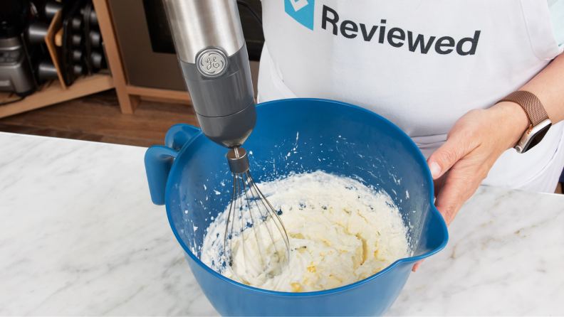 人与围裙站在厨房countertop while using GE Immersion Blender with whisk attachment to whip cream inside of blue bowl.