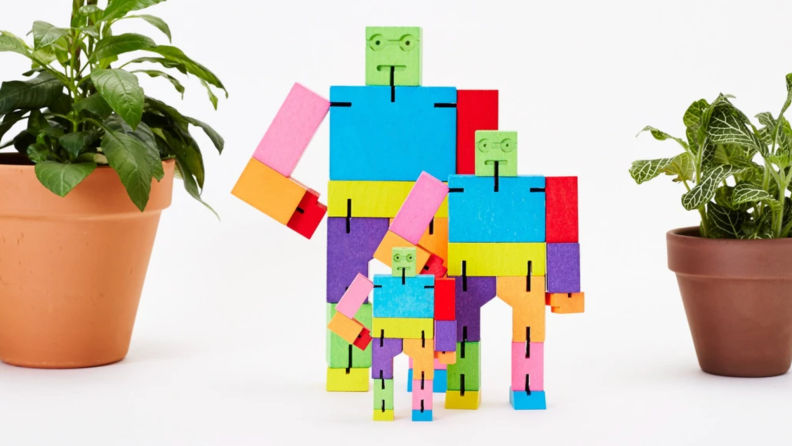 Three different sized multi-colored cube shaped robot toys made of wood in between two green houseplants in clay pots.