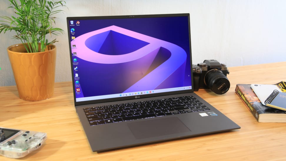 A laptop sits on a desk surrounded by a house plant, video game device, camera, and book.