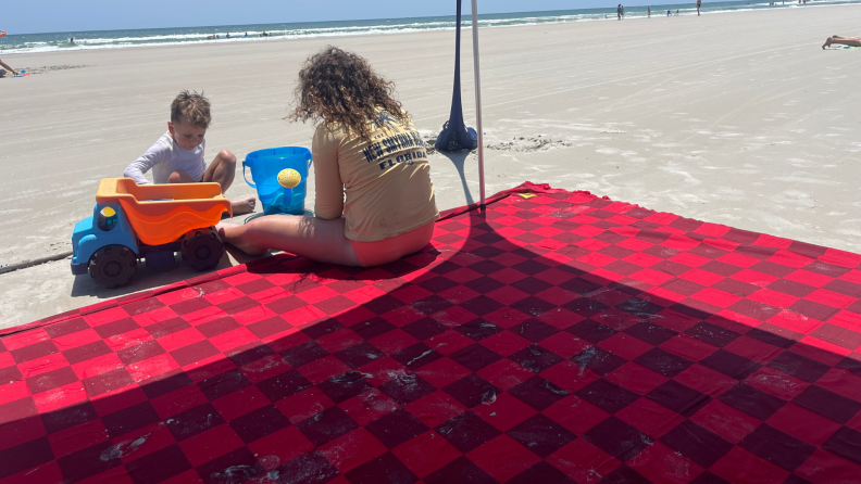 Two small children playing on the edge of red and burgundy Big Beachy blanket on sand at beach.