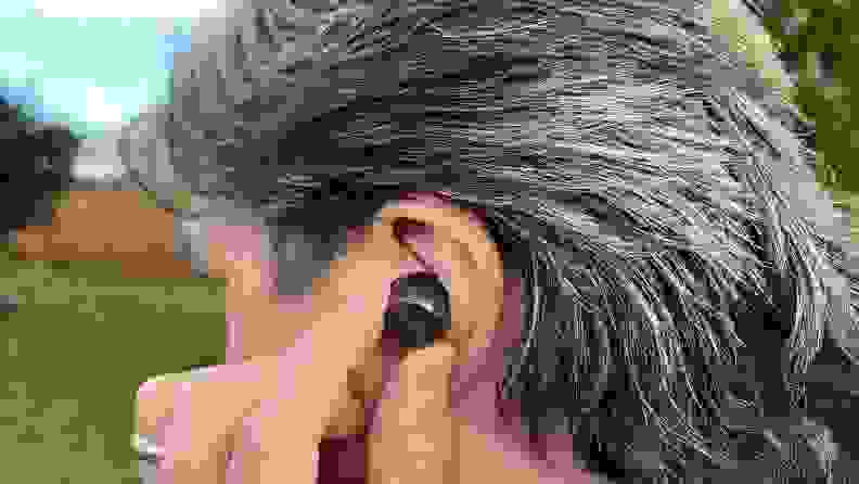 The gunsmoke colored wireless earbud sits in the ear of a man with brown and silver hair, with green grass and blue sky beyond.