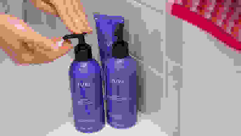A hand reaching to pump a purple bottle of shampoo sitting on the side of a tub with other products.