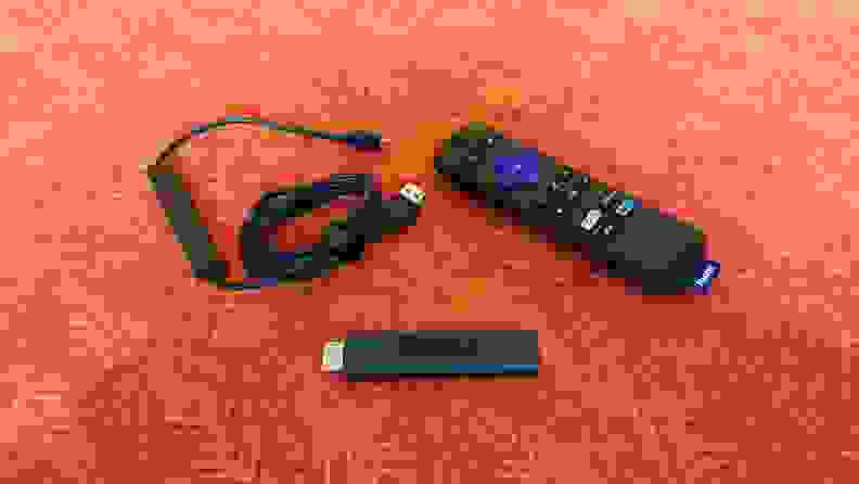 The matte black, candy-bar sized Roku Streaming stick is held with its port showing above a orange-red table with accessories.