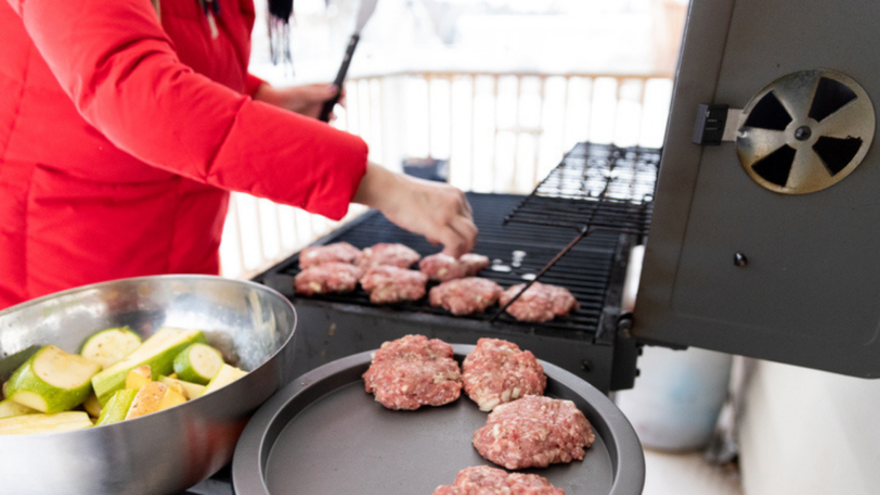 Person in red puffy coat cooking burger patties and vegetables on grill during winter