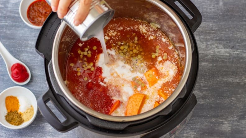 A hand pours ingredients into a pressure cooker that is already partially filled with other ingredients.
