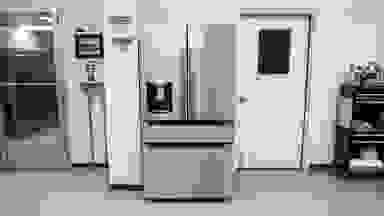 A stainless-steel refrigerator with exterior dispenser stands in a white lab environment next to a white door with a black window.