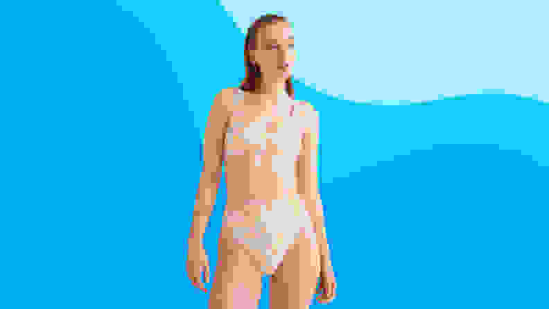 A young blonde woman with damp hair wearing a light pink printed two-piece bathing suit looks off to the right against a patterned blue background