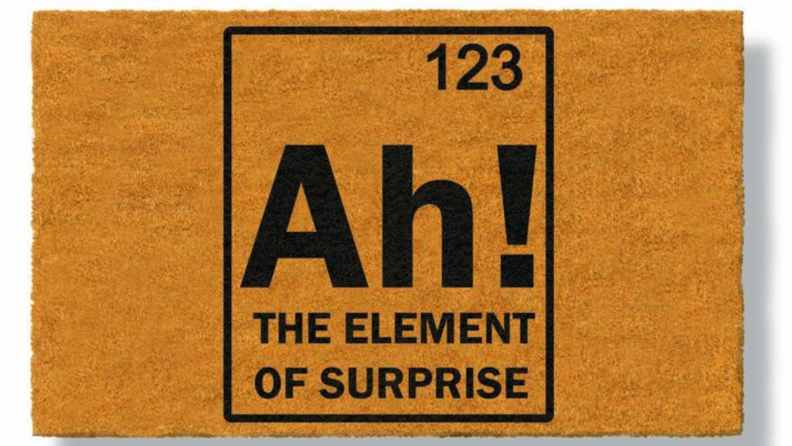 Product shot of tan colored doormat that reads, "Ah! The element of surprise"