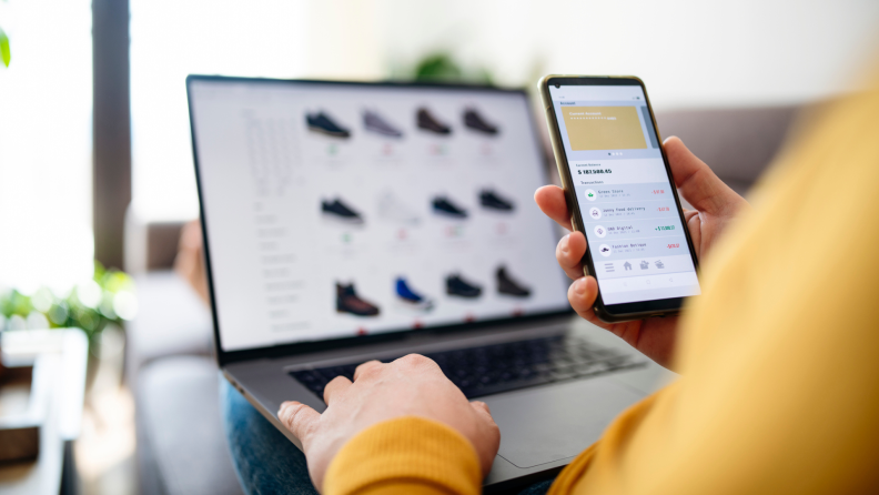 Person sits on couch as they review their online payment plans on smartphone in front of laptop.