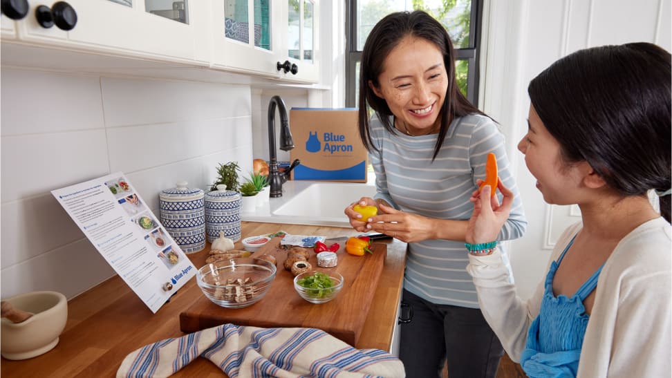 A mother and daughter smile while preparing a meal together in a kitchen with a Blue Apron meal kit.