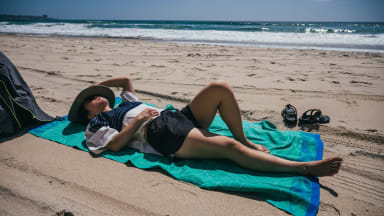 A woman reads a book while laying on a beach towel on the sand.
