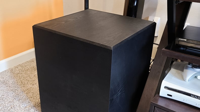 An all black cubed cabinet is shown next to a TV console with a beige and orange wall behind it.