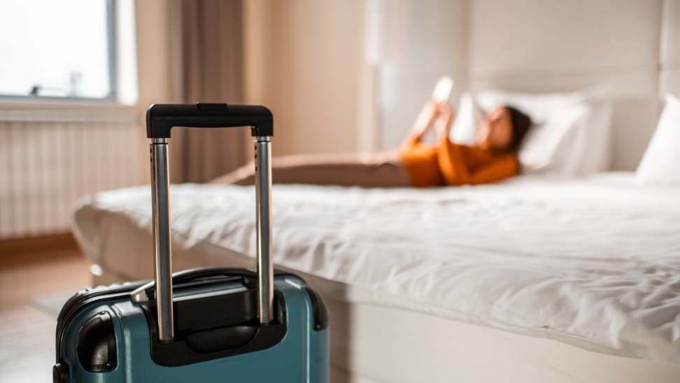 A person is laying on their hotel room bed looking at their phone. A suitcase is at the foot of the bed.