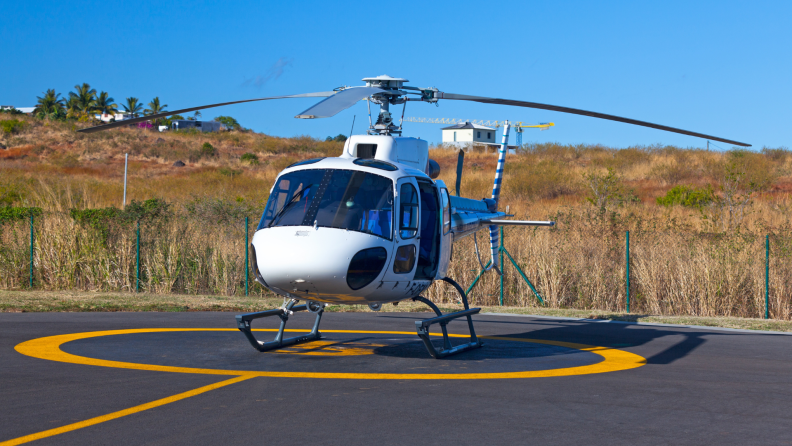 A white helicopter sits on a marked helipad under a clear blue sky, surrounded by dry grass and hills.
