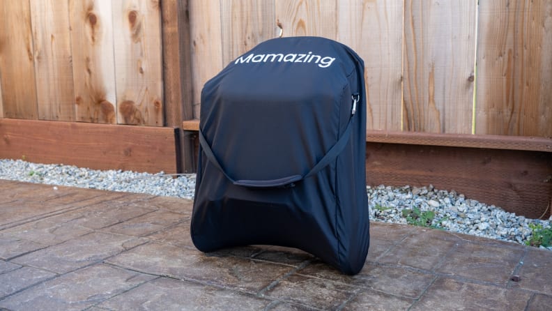 Black Mamazing stroller folded up in its carrying case with the Mamazing logo displayed on the case, in front of a wooden fence sitting on a patio with rocks on the ground behind it.