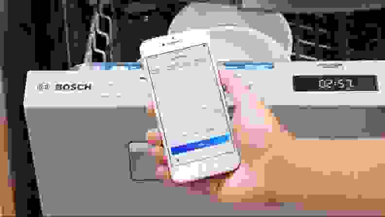 A hand holding up a smartphone displaying the Bosch app, in front of a dishwasher.