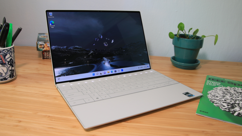 An open silver laptop sits on a desk surrounded by office objects.