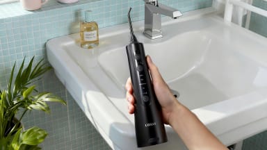 Hand holding a black COSLUS water flosser over a sink next to a potted plant