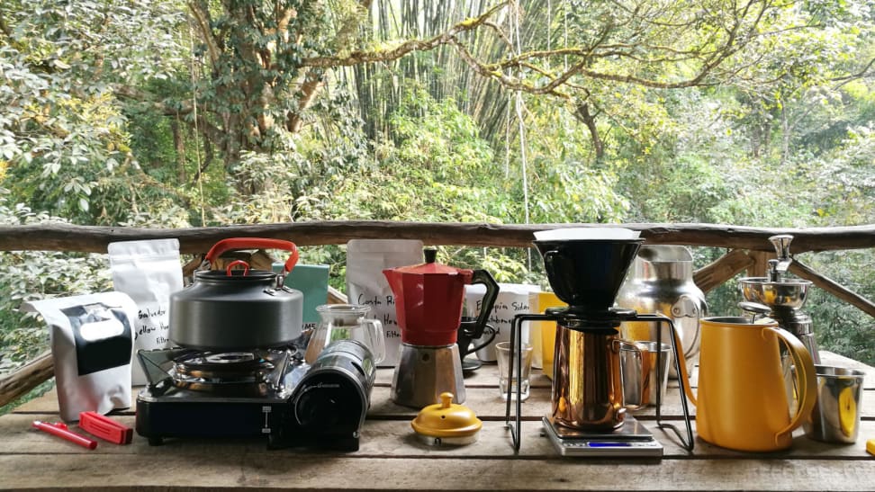 On an outdoor kitchen table, there are some coffee and cooking gadgets. From the left, there's a bag of coffee beans, a stove with a kettle on top, a bialetti moka pot, a pour-over coffee set up, and a milk jug.