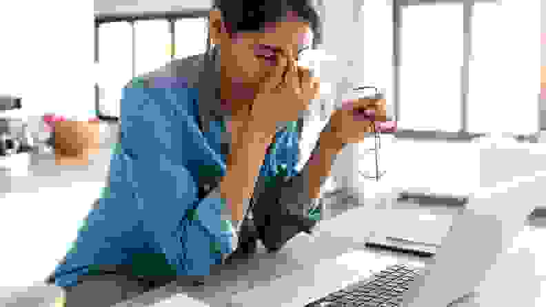 a person rubs their eyes and takes off their glasses sitting by a computer