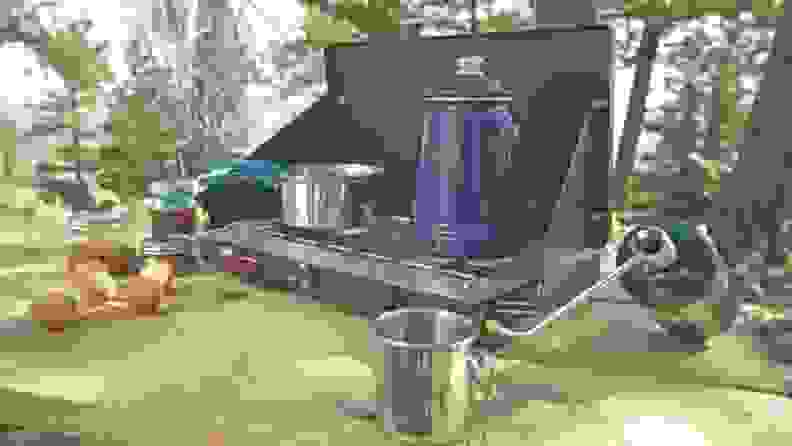 A Coleman two-burner stove is connected to a propane fuel cylinder. On the stove, there's a kettle and a pan.