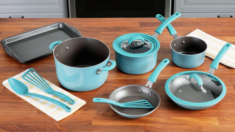 Turquoise blue colored pans, pots, lids and cooking utensils next to small rectangular baking sheet on top of wooden countertop surface.