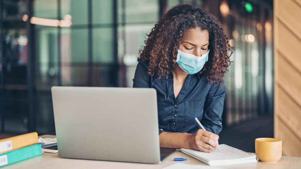 Woman wearing mask writing near open computer