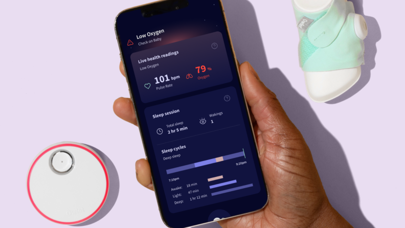Person holding a smartphone with the Owlet app on screen next to a charging doc and Dream Sock.