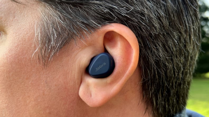 A navy blue earphone sits in the ear of a brown and gray haired man with trees and grass in the background.
