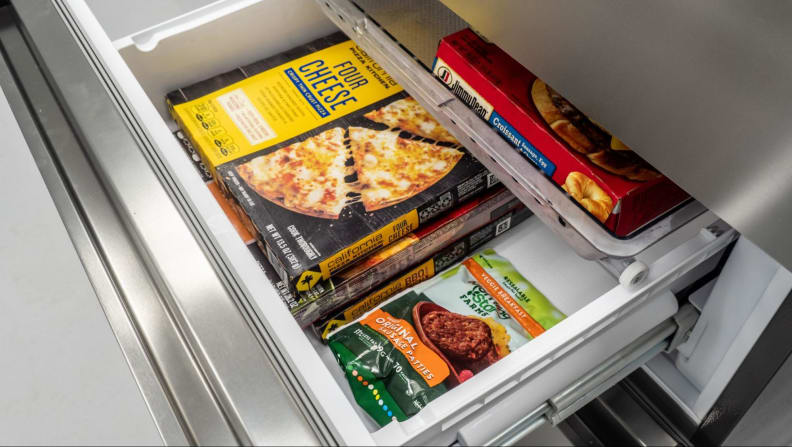 The freezer drawer below is open, showing a box of frozen pizza and some frozen burger patties.