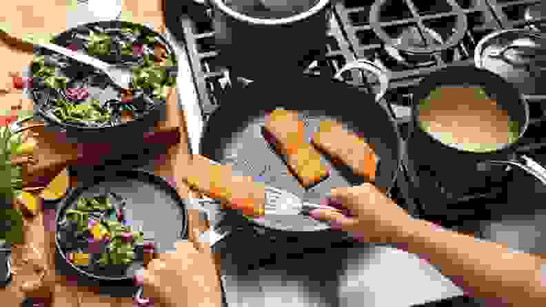 A person cooking three salmon fillets on an Anolon X skillet, surrounded by other Anolon X cookware