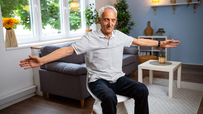 Older person sitting on stool with arms outstretched in a yoga pose.