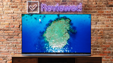 木桌上放着一台OLED电视，屏幕上有一个岛，前面是一面砖墙，墙上挂着霓虹灯，上面写着“已审核”。