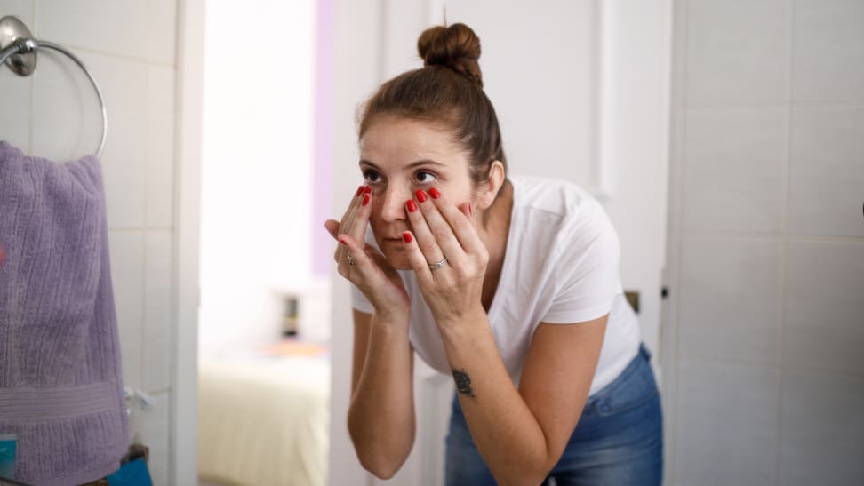 A woman hunched over in the mirror with her hands under her eyes.