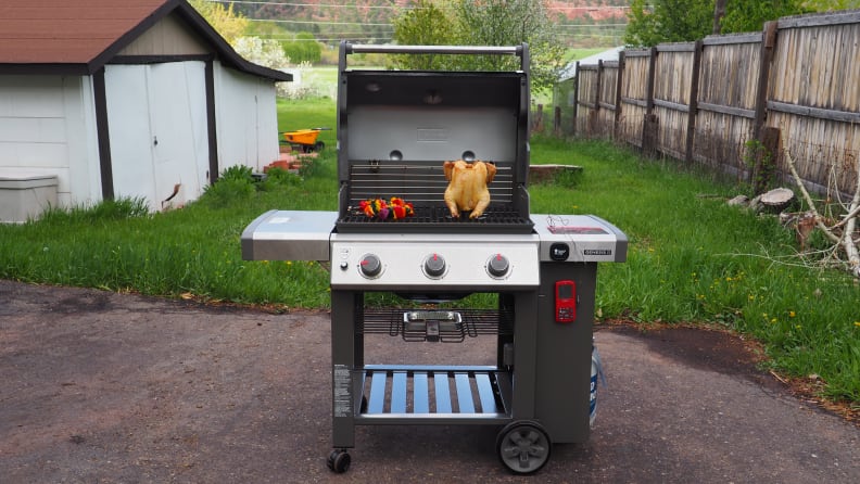 The Weber Genesis II E-310 open, revealing chicken and vegetables grilling, in a backyard setting