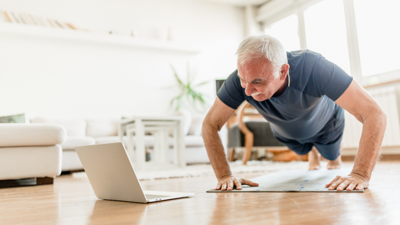 A person does push-ups at home next to an open laptop.