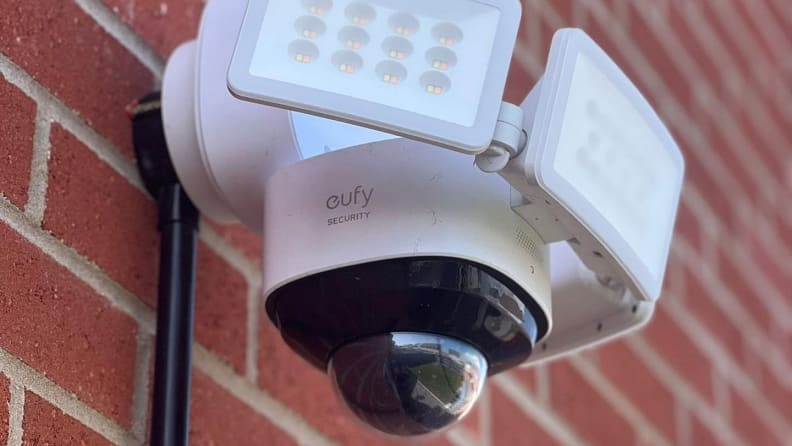 White surveillance system with built-in cameras and lights mounted on brick wall.