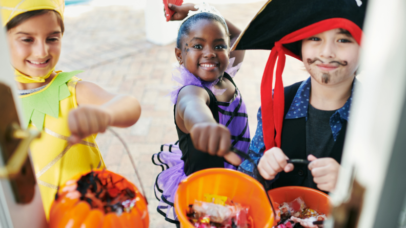 Three small children in Halloween costumes stand at a door and hold out candy buckets.