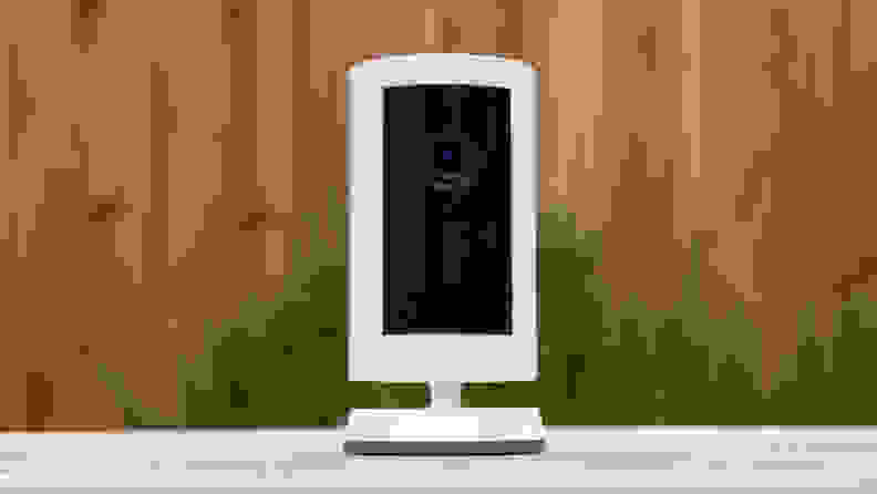 A white Ring camera stands on a white table with a brown fence in the background