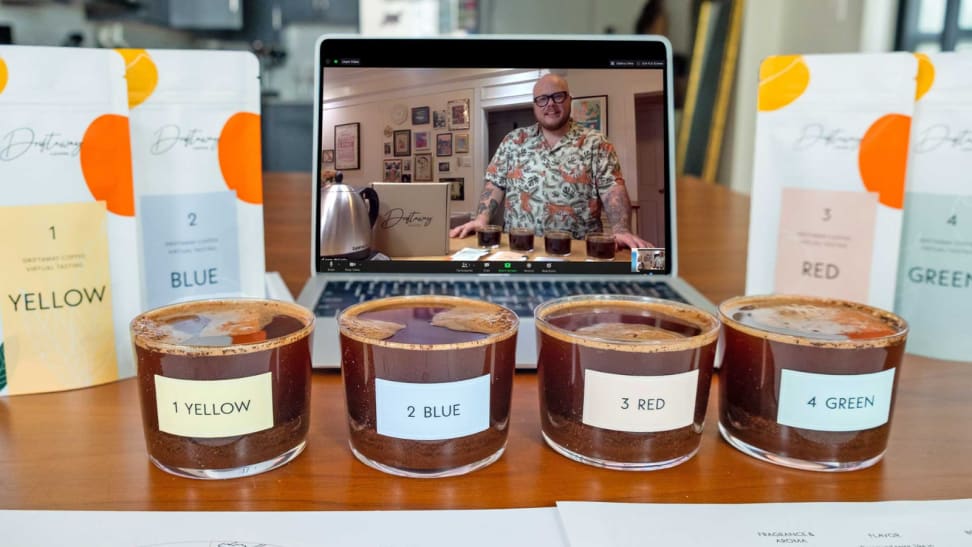 A virtual coffee tasting setup including a laptop streaming the class, clear cupping glasses with colored labels filled with the different coffees, and the corresponding colored coffee bean bags from Driftaway.