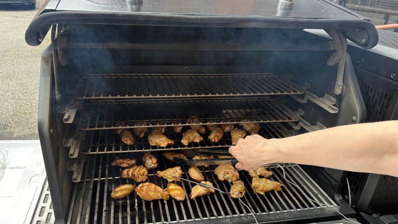 Person grabbing chicken wing with tongs from an open grill filled with smoked chicken wings