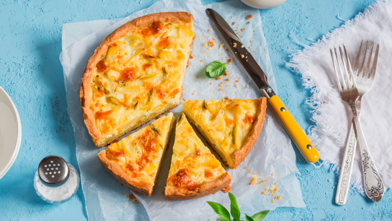 A sliced quiche on a blue surface, surrounded by utensils.