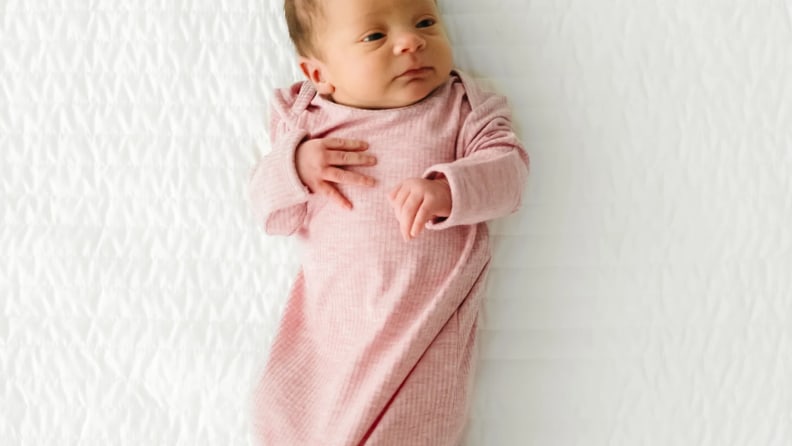 Newborn baby in a pink gown lying awake on a white textured blanket.